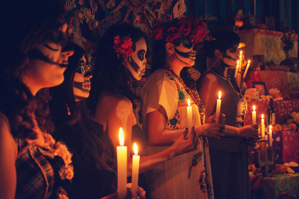 Catrinas frente al altar del Día de Muertos portando velas.